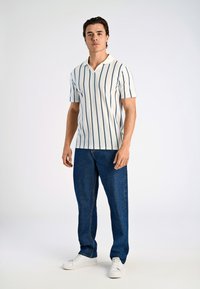 Vit polo skjorta med blå vertikala ränder, korta ärmar och V-ringad krage, matchad med blå denimjeans och vita sneakers.