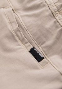 Pantalon beige au toucher lisse, avec une poche latérale et une étiquette de marque noire avec un texte blanc cousu dans le tissu.