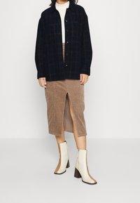 Camicia a quadri blu navy con bottoni, gonna midi in corduroy beige con spacco frontale e stivali a blocco con tacco color crema e dettagli marroni.