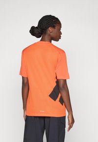 Oranž lühikeste varrukatega võrkpoliester spordisärk mustade kangakasutustega seljal, ümmarguse kaeluse ja mugava lõikega.