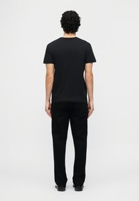 Άνδρας με σγουρά μαλλιά, όρθιος και γυρισμένος με την πλάτη, φοράει απλό μαύρο μπλουζάκι, μαύρο παντελόνι και μαύρα παπούτσια σε λευκό φόντο.