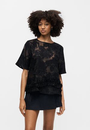 Γυναίκα με κατσαρά μαλλιά που φοράει κοντομάνικη μαύρη διάφανη μπλούζα με λουλούδια και μαύρη μίνι φούστα, στέκεται μπροστά σε καθαρό λευκό φόντο.