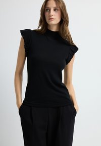 Zwart, nauwsluitend topje met een hoge hals en gerimpelde afgezette mouwtjes. Gladde textuur, gecombineerd met op maat gemaakte zwarte broek. Eenvoudig en elegant ontwerp.