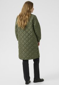 Quilted lang olivengrøn jakke med en glat tekstur, der har en bred halsudskæring og sidelommer. Sammen med sorte bukser og sko.