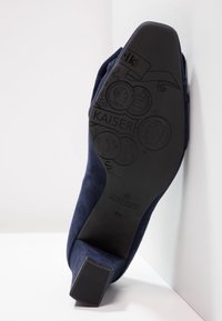 Scarpa in suede blu navy con suola texture, caratterizzata da un tacco quadrato e incisioni decorative di monete e branding sul fondo.