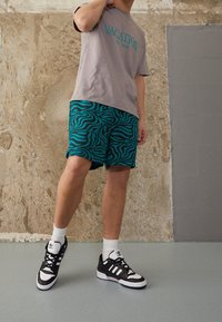 Chemise grise à manches courtes avec le texte "VACATION", short zébré vert et noir, chaussettes blanches et baskets noires avec accents blancs.
