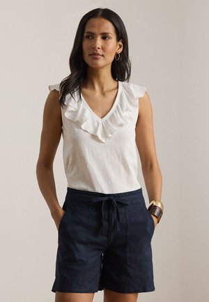 Γυναίκα που φοράει λευκό μπλουζάκι χωρίς μανίκια με βολάν στο λαιμό, σκούρο μπλε σορτς με κορδόνι, χρυσούς κρίκους στα αυτιά και φαρδύ βραχιόλι, στέκεται με τα χέρια στις τσέπες.