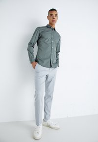 Camisa de botones gris claro, pantalones gris claro entallados y zapatillas blancas con detalles de cinta; entorno interior con fondo neutro.