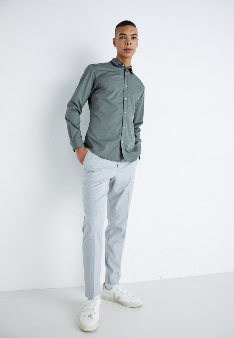 Helles graues Hemd mit Knopfleiste, taillierte hellgraue Hose und weiße Sneaker mit Riemendetails; Innenraum mit neutralem Hintergrund.