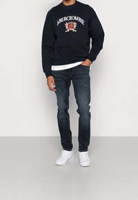 Námořnická mikina s logem "Abercrombie", bílými detaily, v kombinaci s tmavými džíny a bílými teniskami. Ležérní outfit na neutrálním pozadí.