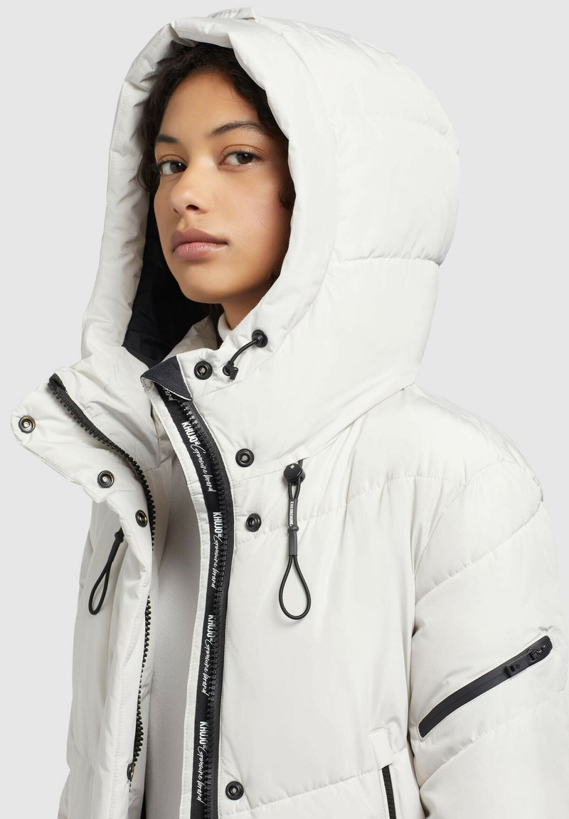 Weißer Pufferjacke mit Kapuze, ausgestattet mit einem Reißverschluss vorne, schwarzen Akzenten, Kordelzügen und seitlichen Taschen. Strukturiertes, gestepptes Design.