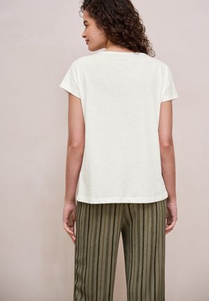 Kobieta z kręconymi włosami, ubrana w krótkim rękawem biały top i oliwkowo-zielone spodnie z wysokim stanem z pionowymi czarnymi paskami, zwrócona tyłem.