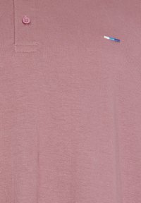 Gros plan sur un polo mauve avec patte de boutonnage à deux boutons et petit logo horizontal brodé en bleu, blanc et turquoise sur la poitrine.