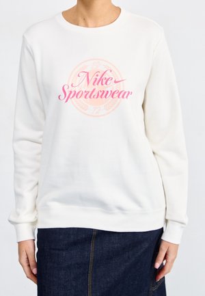 Kvinde iført en hvid langærmet sweatshirt med pink "Nike Sportswear" tekst og et cirkulært emblem, kombineret med en mørkeblå nederdel.