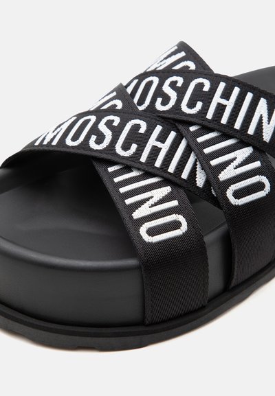 Sandale plateforme noire avec des larges brides croisées présentant un texte blanc "MOSCHINO" en lettres grasses.