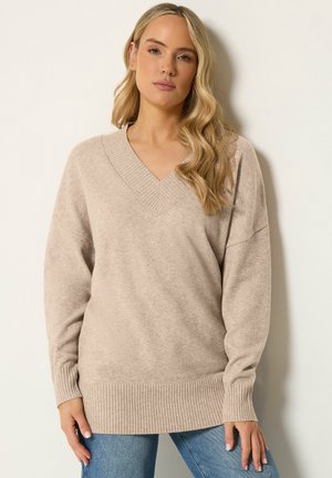 Frau mit langen blonden Haaren, die einen beigen Pullover mit V-Ausschnitt und blaue Jeans trägt, steht vor einem hellen Hintergrund.