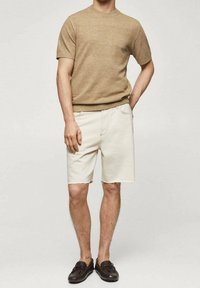 Pull à manches courtes beige en maille associé à un short en denim blanc. Le short présente un ourlet effiloché et des poches. Le mannequin porte des chaussures d'été à enfiler de couleur marron foncé.
