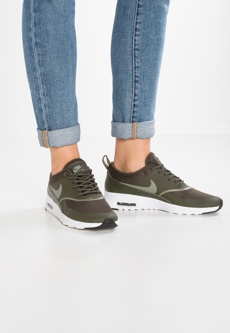 Nohy v olivově zelených teniskách Nike Air Max Thea s bílou podrážkou a vyhrnutými modrými džíny na bílém podlaze.