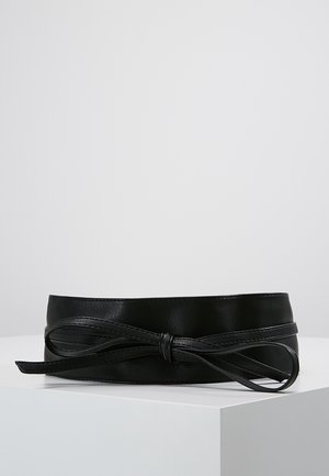 Ceinture large en cuir noir avec une fine lanière nouée en un nœud à l'avant, présentée sur un piédestal blanc sur un fond gris clair.