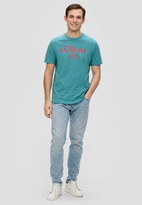 Turquoise t-shirt met rode print, ronde hals, gecombineerd met lichtblauwe jeans en witte sneakers. Soepele katoentextuur. Casual fit.