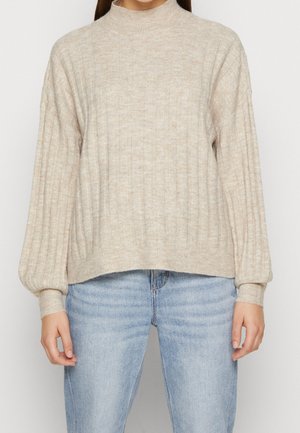 Femme portant un pull en maille côtelée beige à manches ballon, associé à un jean en denim bleu clair, debout devant un fond uni.