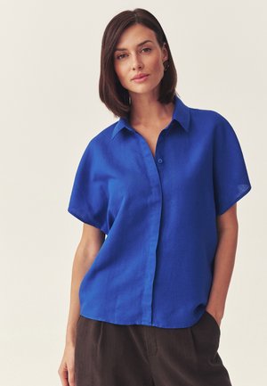 Kobieta o ciemnych włosach do ramion, ubrana w luźną, jaskrawo niebieską koszulę na krótki rękaw na guziki oraz ciemne spodnie, stojąca na jednolitym tle.