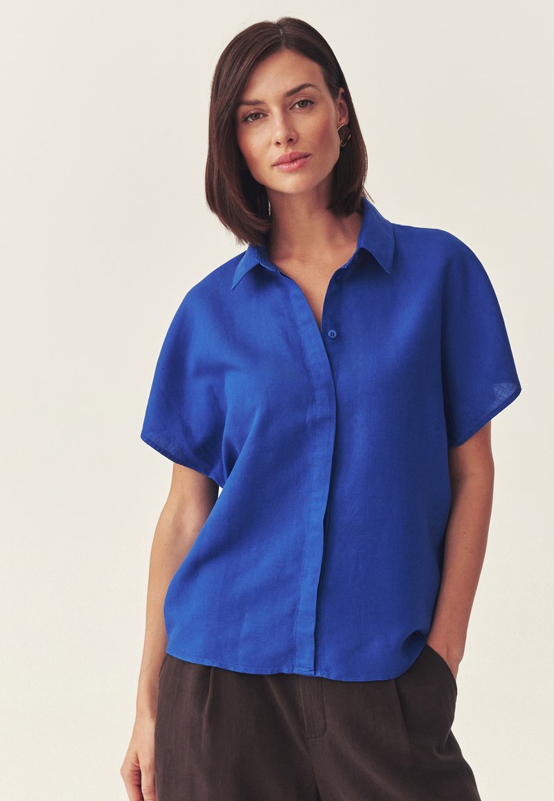 Femme aux cheveux bruns mi-longs portant une chemise ample à manches courtes boutonnée de couleur bleu vif et un pantalon foncé, debout devant un fond uni.