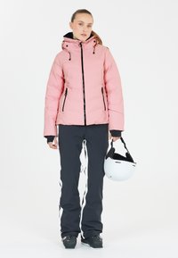 Rosa vadderad skidjacka med svarta detaljer, kombinerad med svarta skidbyxor med vita ränder och en vit skidhjälm i handen.