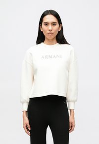 Λευκή φούτερ με τη λέξη "ARMANI" σε λαμπερά γράμματα, στρογγυλή λαιμόκοψη και χαμηλωμένους ώμους. Συνδυασμένη με μαύρο ριμπ παντελόνι.