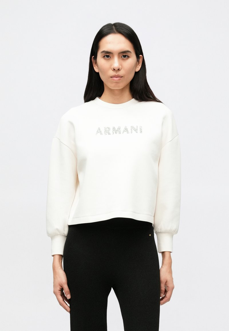 Λευκή φούτερ με τη λέξη "ARMANI" σε λαμπερά γράμματα, στρογγυλή λαιμόκοψη και χαμηλωμένους ώμους. Σ υνδυασμένη με μαύρο ριμπ παντελόνι.