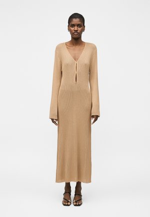 Kobieta stojąca, ubrana w długą beżową prążkowaną sukienkę z dzianiny z długimi rękawami, z detalami w formie otworu przy głębokim dekolcie w kształcie litery V oraz czarnymi sandałami na paski.