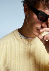 Gehaakte gele trui met een ribbelhalslijn, over een wit shirt. Zwarte zonnebril en een zilveren kettingaccentueren de look.