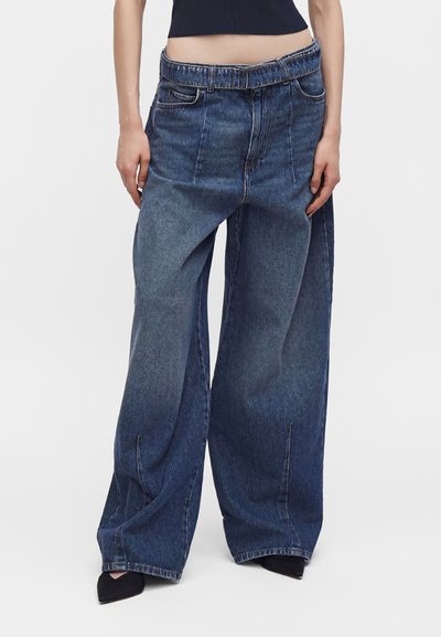 Personne portant un jean en denim bleu à larges jambes surdimensionnées avec une taille haute et des coutures devant, assorti à des chaussures noires pointues et un top court.