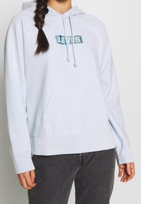 Sudadera con capucha azul claro hecha de tela suave, con bolsillo frontal, capucha ajustable con cordón y un logotipo verde de Levi's en el pecho. Diseño sencillo y casual.