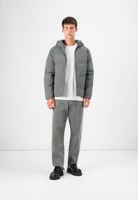 Grijze geïsoleerde pufferjack met een capuchon, met een ritssluiting aan de voorkant, grote opgestikte zakken, gedragen over een wit t-shirt en grijze jeans. Zwarte schoenen.