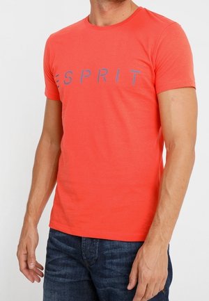 Mann trägt ein körpernahes korallenrotes Esprit-T-Shirt mit blauem Logo und dunkelblaue Jeans, steht mit entspannten Armen.