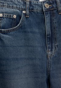 Gros plan d'un jean en denim bleu montrant la poche avant, la passants de ceinture, le bouton en métal et les détails de couture.