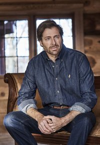 Jeanshemd in tiefem Blau mit langen Ärmeln, durchgehender Knopfleiste und Logo auf der Brust. Kombiniert mit passenden blauen Jeans. Hintergrund mit Holzvertäfelung.