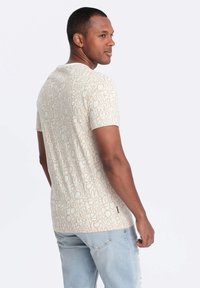 Vaftet beige t-skjorte med kort erme og et gjentatt tekstmønster i lysebrune toner. Den har rund hals og en avslappet passform, og er brukt sammen med lyseblå jeans.