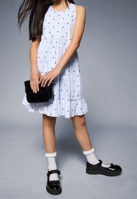 Bílé šaty s černým vzorem srdcí, rozšířeným lemem, kombinované s černými lakovanými botami Mary Jane a bílými krajkovými ponožkami. Černá kabelka přes rameno.