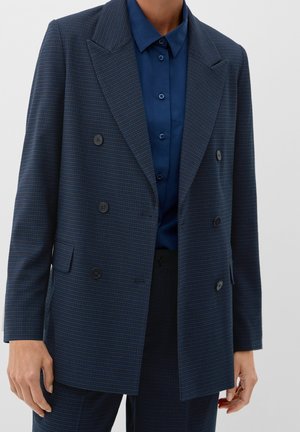 Person trägt einen dunkelblauen karierten zweireihigen Blazer über einem geknöpften marineblauen Hemd und passenden dunklen Hosen.