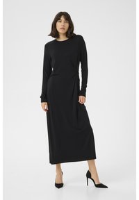 Long black jersey dress with a twist detail at the waist, round neckline, and long sleeves; paired with black stiletto heels.