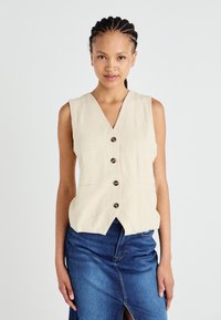 Beige ärmellose Weste mit V-Ausschnitt, fünf dunklen Knöpfen, zwei Fronttaschen und einem leicht abgerundeten Saum. Getragen über einem Jeansrock.