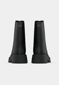 Μαύρες μπότες αστραγάλου από μαλακό συνθετικό υλικό, με ψηλό κορμό, πίσω φερμουάρ και χοντρές ελαστικές σόλες με κανάλια πρόσφυσης.