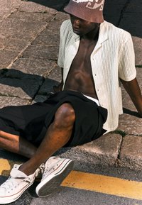 Witte gestructureerde korte mouwen overhemd, zwarte shorts en witte sneakers. De persoon zit op stenen bestrating, met een gebreide beige bucket hat.