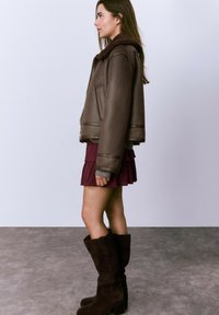 Chaqueta de cuero marrón cortada con cuello de piel, combinada con una falda borgoña plisada y altas botas de ante marrón.