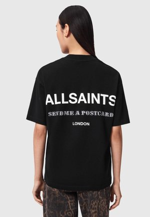 Kvinde med langt mørkt hår set bagfra iført sort T-shirt med hvid tekst "ALLSAINTS SEND ME A POSTCARD LONDON" og mønstrede bukser.