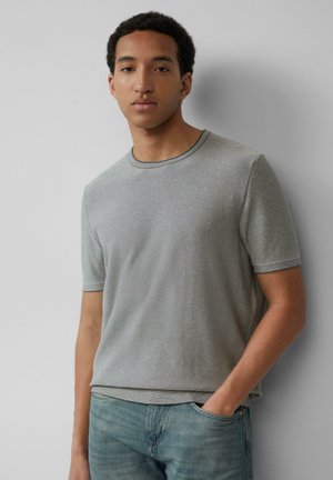 T-shirt gris à manches courtes avec un col rond côtelé, présentant un motif à rayures subtil. Porté avec un jean bleu clair, les mains dans les poches.