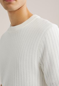 Close-up van een persoon die een wit gebreide trui met structuur draagt met een geribde ronde hals, tegen een effen achtergrond.