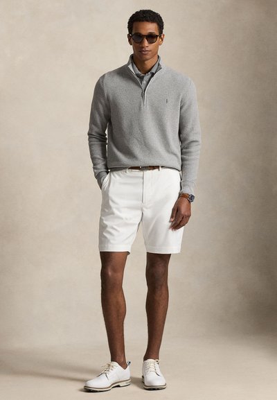Uomo che indossa occhiali da sole, maglione grigio con cerniera a metà, polo a righe, pantaloncini bianchi, cintura marrone, orologio e scarpe bianche con lacci, in piedi con le mani in tasca.